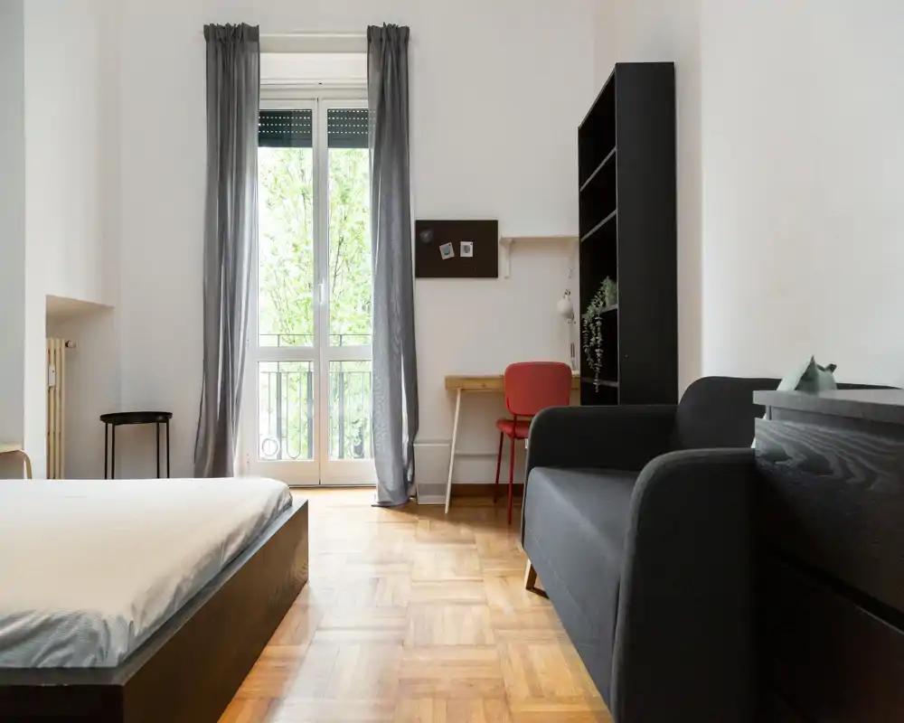 Camera da letto luminosa e moderna con letto basso, ampia finestra con tende, piccolo angolo studio accanto a una libreria e divanetto.