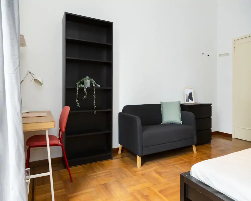 Angolo di camera luminoso e ordinato con un divano due posti, una libreria aperta, una scrivania con sedia e parte del letto visibile in primo piano. Pavimenti in parquet caldo e arredo minimale, ideale per affitti compatti.