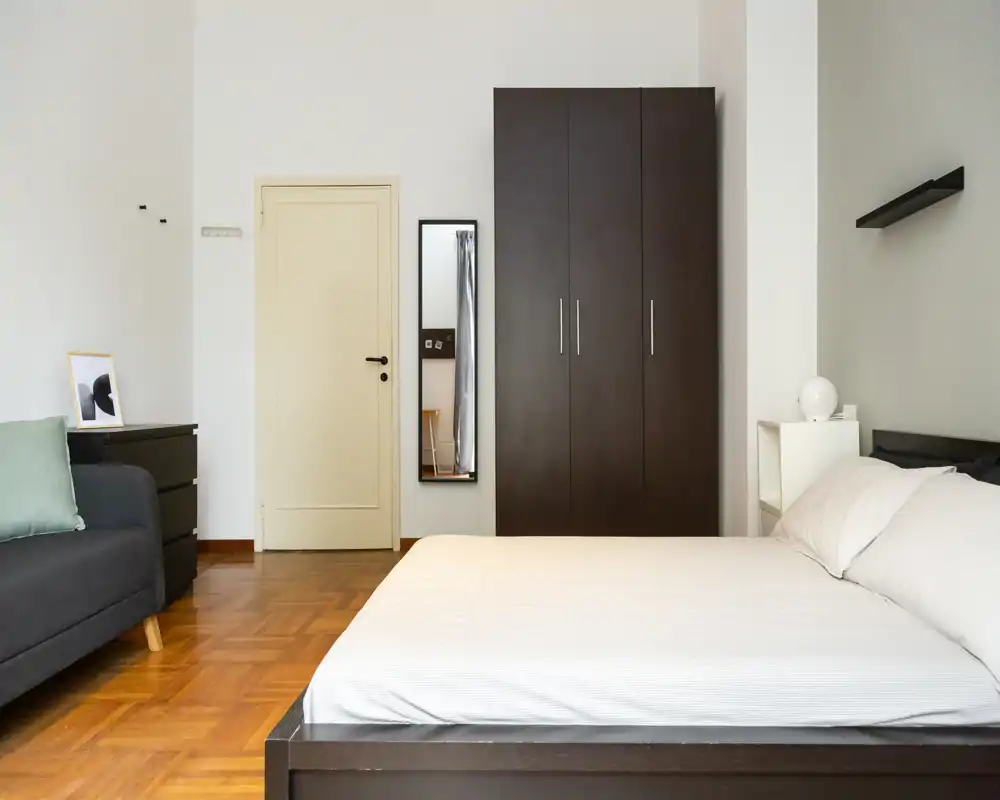 Camera da letto luminosa e moderna con un grande letto in primo piano, un armadio scuro e un piccolo divano. Linee pulite, parquet in legno e arredo minimal creano un'atmosfera tranquilla e confortevole.