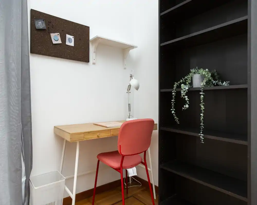 Angolo living/lavoro compatto e ben arredato con scrivania in legno, sedia rossa e scaffale aperto; luminoso e ordinato, ideale per smart working o lettura.