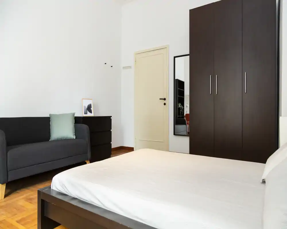 Camera da letto luminosa e minimal con letto matrimoniale ben rifatto, armadio in legno scuro e un divanetto che crea un angolo accogliente.