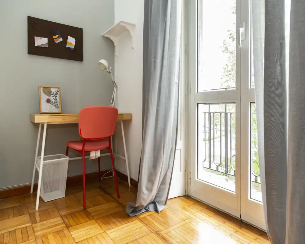 Rincón luminoso y minimalista con un escritorio pequeño y una silla roja junto a una gran ventana/puerta — ideal como zona de trabajo compacta con luz natural.