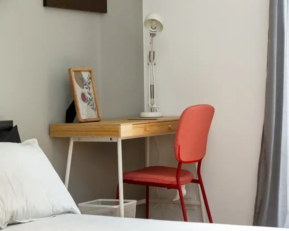 Rincón de dormitorio luminoso y minimalista con el borde de la cama visible, escritorio de madera, lámpara y una silla coral: un espacio de trabajo compacto dentro del dormitorio.