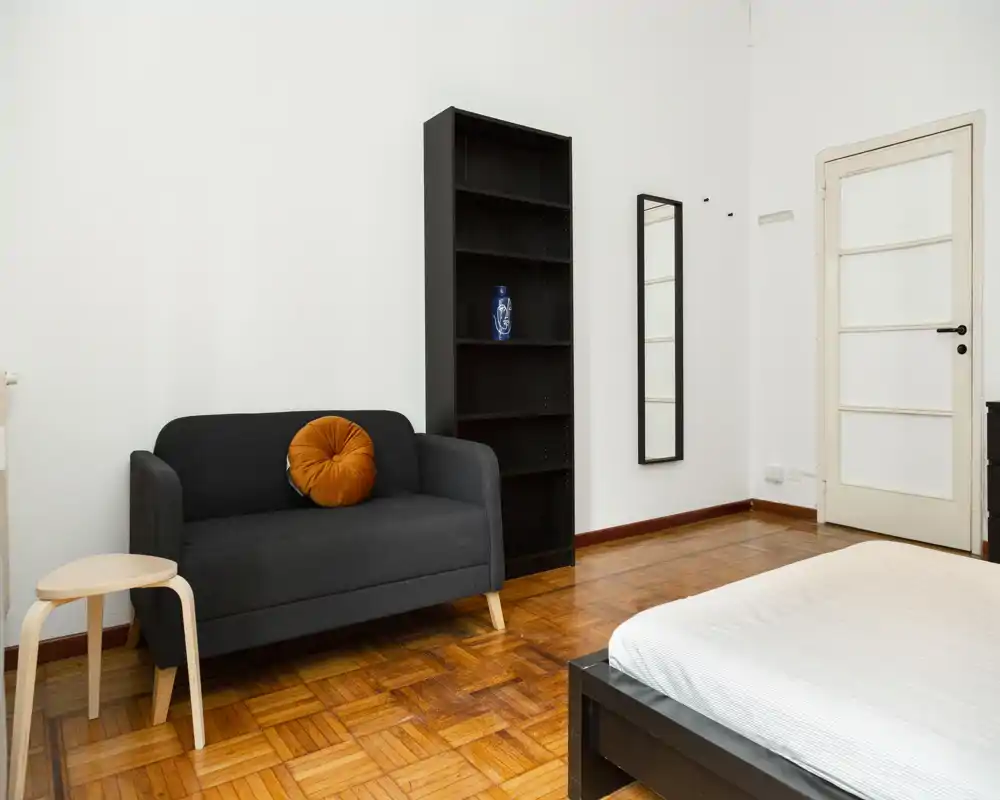 Dormitorio luminoso y de mobiliario sencillo: cama visible, pequeño sofá con cojín decorativo, estantería y espejo de cuerpo entero — ideal para un anuncio minimalista.
