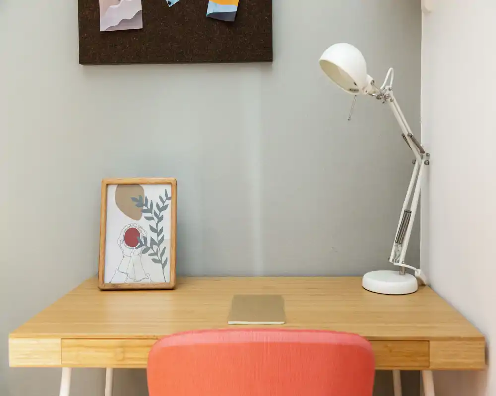 Rincón de escritorio luminoso y minimalista con mesa de madera, lámpara blanca ajustable y silla coral — un acogedor espacio de trabajo.