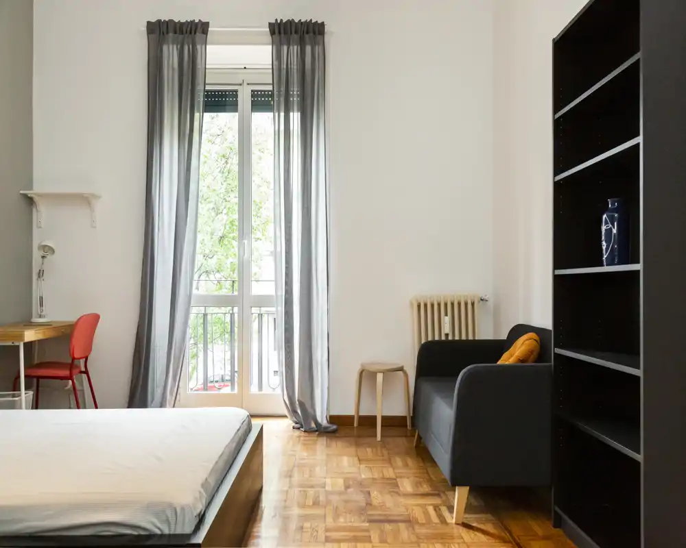 Dormitorio luminoso y minimalista con cama baja, gran ventana con cortinas traslúcidas, pequeño escritorio con silla y sofá compacto, ideal para una persona o pareja que busque un espacio aireado.