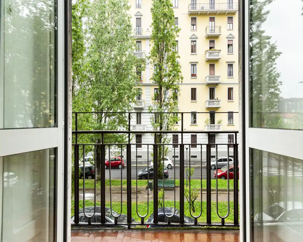 Balcón enmarcado por puertas de vidrio abiertas, con vistas a una calle arbolada y la fachada de un edificio residencial clásico.