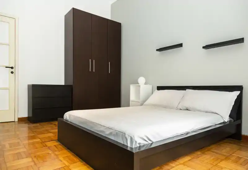 Dormitorio luminoso y minimalista con cama doble baja, ropa de cama blanca y armario de madera oscura. Líneas limpias, suelo de parquet y estantes sencillos crean un ambiente moderno y tranquilo, ideal para anuncios de alquiler.