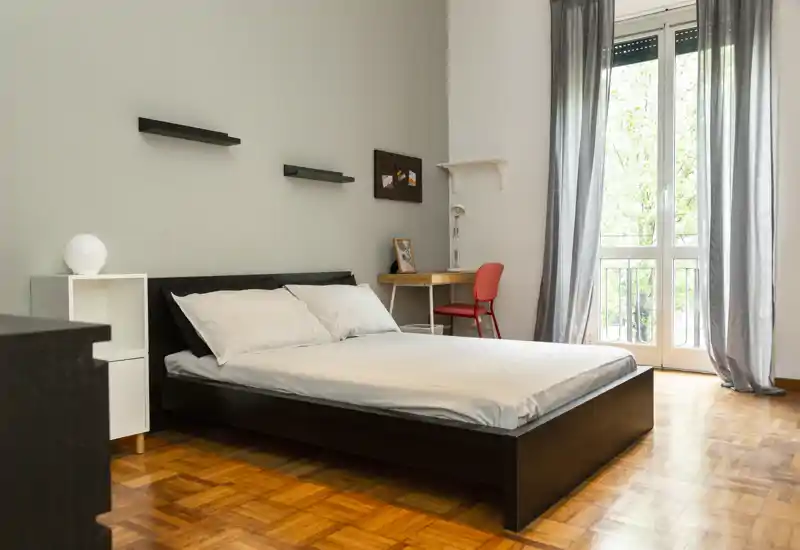 Dormitorio luminoso y minimalista con cama baja doble, suelo de parquet y una ventana que aporta mucha luz natural. Un pequeño escritorio y una silla junto a la ventana ofrecen un espacio de trabajo funcional.
