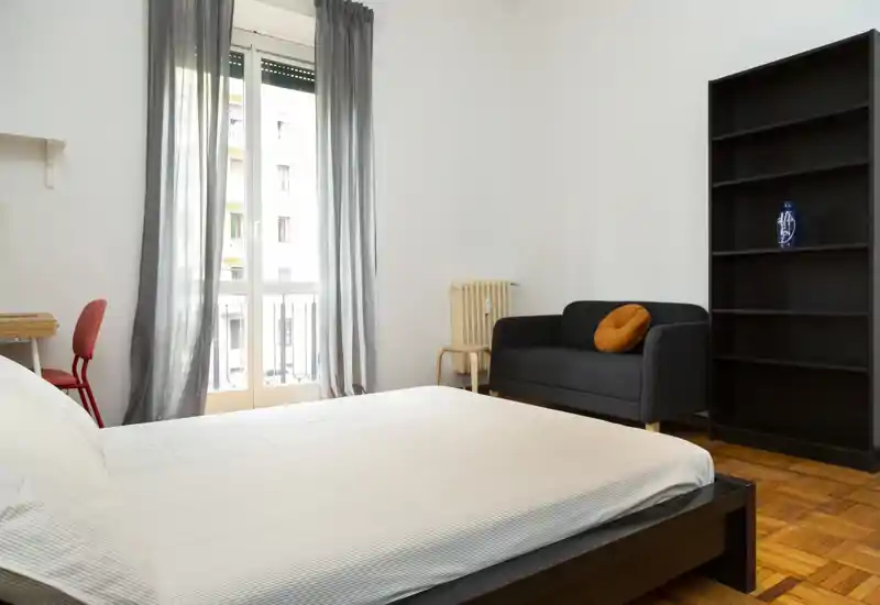 Dormitorio luminoso y minimalista con una cama grande en primer plano, un pequeño sofá y estantería. Suelo de parquet y una ventana alta con cortinas translúcidas que aportan luz natural suave.