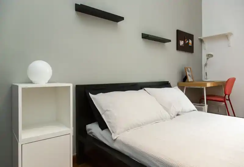 Dormitorio minimalista y luminoso con cama doble, ropa blanca, mesita tipo cubo y un pequeño escritorio con silla. Estilo limpio y moderno, adecuado para alquileres temporales o estudiantes.
