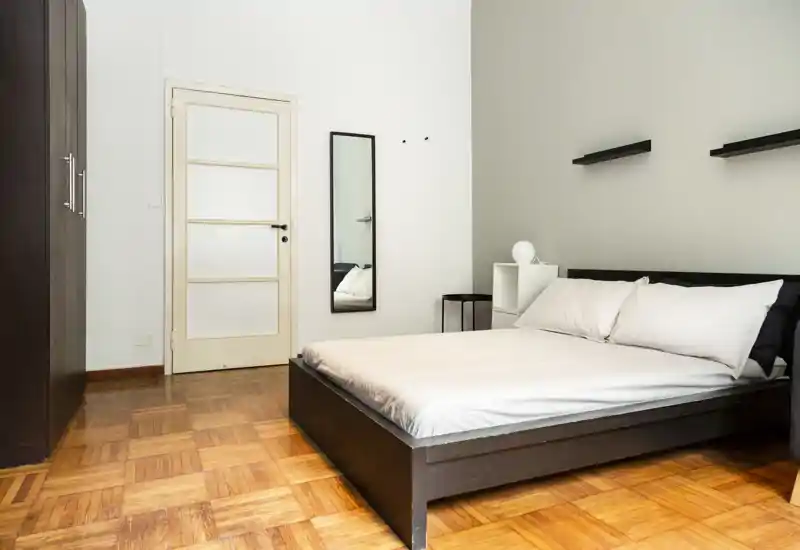 Camera matrimoniale minimalista con letto basso in legno scuro, biancheria neutra, piccola mensola/comodino e specchio a tutta altezza — ambiente luminoso, pulito e arredato in stile semplice, ideale per annunci.