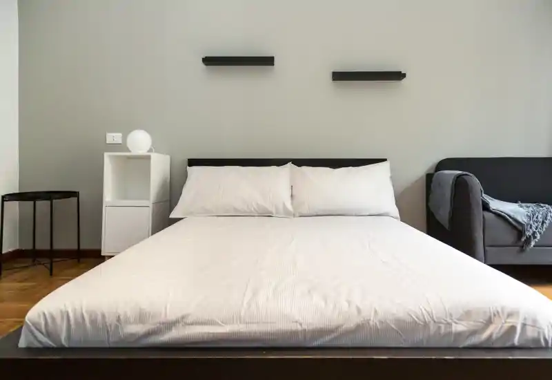 Camera da letto luminosa e minimale con un ampio letto inquadrato centralmente, biancheria bianca, un piccolo ripiano e un divano visibile a destra — perfetta per annunci che valorizzano uno spazio notte pulito e moderno.