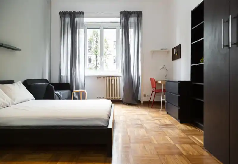 Camera da letto luminosa e moderna con ampio letto davanti alla finestra, parquet, un piccolo divano e una scrivania—ben illuminata e arredata con semplicità.