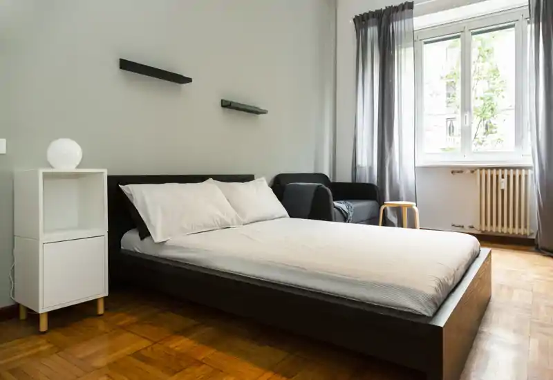 Camera da letto moderna e luminosa con letto matrimoniale basso, comodino e una poltroncina vicino alla finestra. Pavimento in legno chiaro e toni grigio chiaro per un’atmosfera minimal e rilassante.