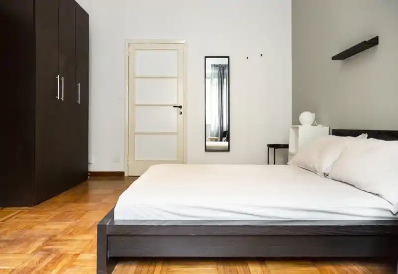 Camera da letto minimalista con letto basso in legno scuro, biancheria bianca e un ampio armadio. Luce naturale morbida e arredo essenziale creano un ambiente moderno e rilassante, perfetto per gli annunci.
