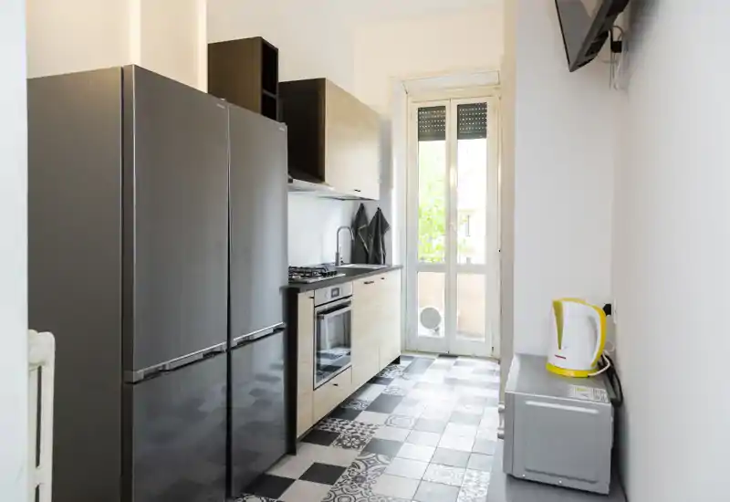 Modern galley kitchen with wood-effect cabinets, built-in oven and gas hob, large dark fridge and patterned tiled floor opening onto a balcony door — bright and well-equipped.