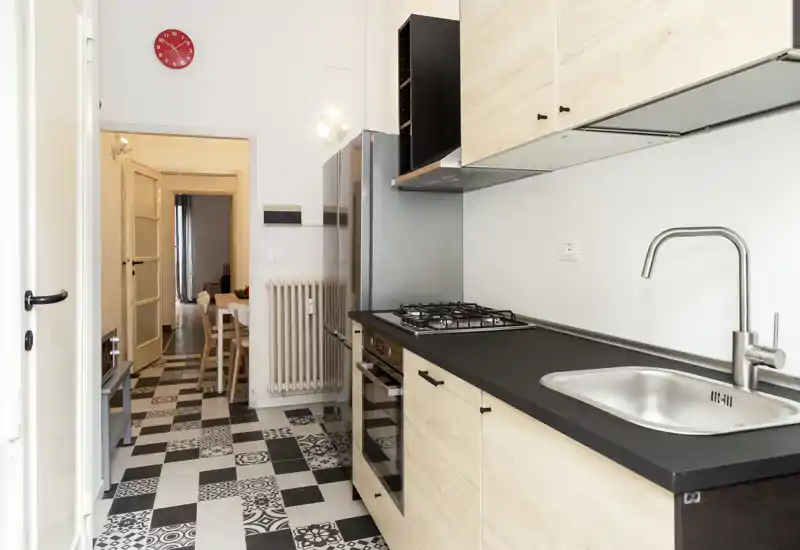Modern galley kitchen with a black countertop, stainless sink and gas hob; light wood cabinets and patterned black-and-white floor tiles create a stylish, functional cooking space.