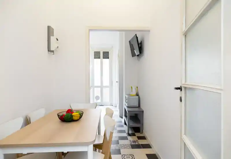 Bright, minimal dining area with a light wood table, white chairs and patterned tile floor, lit by natural light from a balcony door.