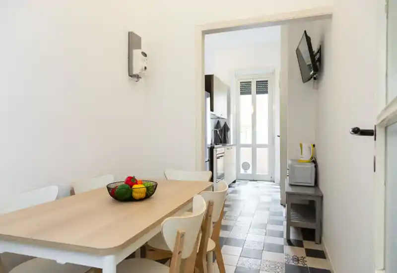 Bright, modern dining area with a light wood table and white chairs, decorative fruit bowl centerpiece and a clear view into the adjacent kitchen and balcony door.