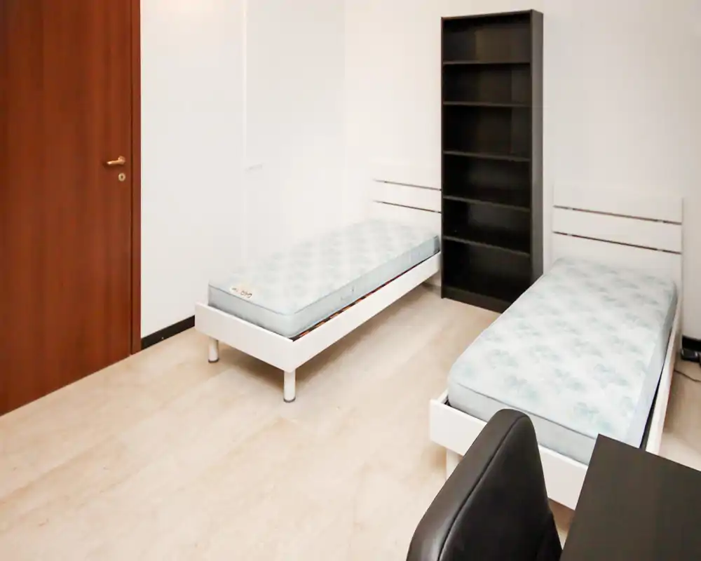Dormitorio luminoso con dos camas individuales de estructura blanca y una estantería alta negra; decoración mínima, ideal para estudiantes o alquileres económicos.