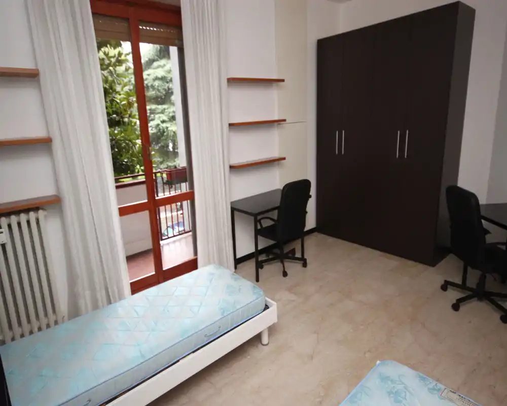 Dos camas individuales en un dormitorio luminoso y amueblado de forma sencilla con puerta a un pequeño balcón, armario y escritorios. La luz natural y tonos neutros lo hacen ideal para estudiantes o alquileres temporales.