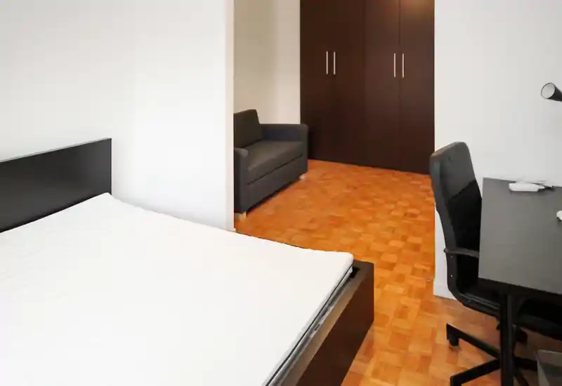 Camera da letto moderna e luminosa con letto matrimoniale visibile, scrivania e sedia, armadio e un piccolo divano nell'angolo — ideale per single o coppie.