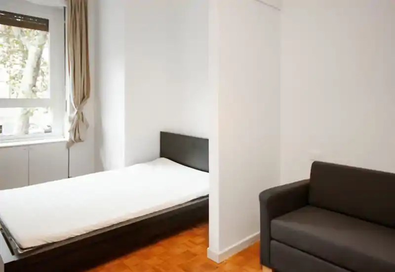 Camera da letto luminosa e minimale con un letto singolo vicino alla finestra e un piccolo divano. Pareti bianche e luce naturale creano un ambiente fresco e arioso, perfetto per un affitto compatto.