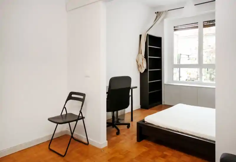 Camera da letto luminosa e minimale con letto basso su piattaforma, scrivania e scaffale accanto a una grande finestra che fa entrare molta luce naturale. Arredamento semplice, ideale per una persona.