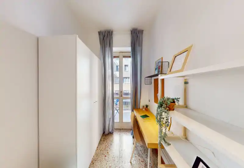 Espacio estrecho tipo dormitorio con un armario alto a la izquierda y un escritorio de madera junto a la puerta del balcón; luz natural brillante y decoración sencilla y funcional.