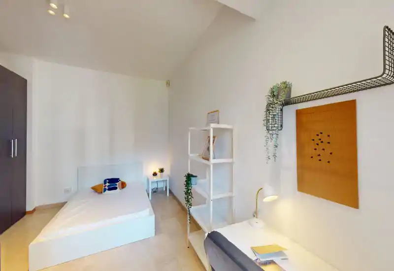 Bright, minimal bedroom with a single bed, side table and shelving. Soft decorative touches include cushions, a desk lamp and hanging plants for a cozy atmosphere.