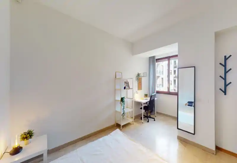 Bright, minimal bedroom with a visible bed in the foreground, a study desk by the window, and simple shelving and mirror — ideal for students or professionals.