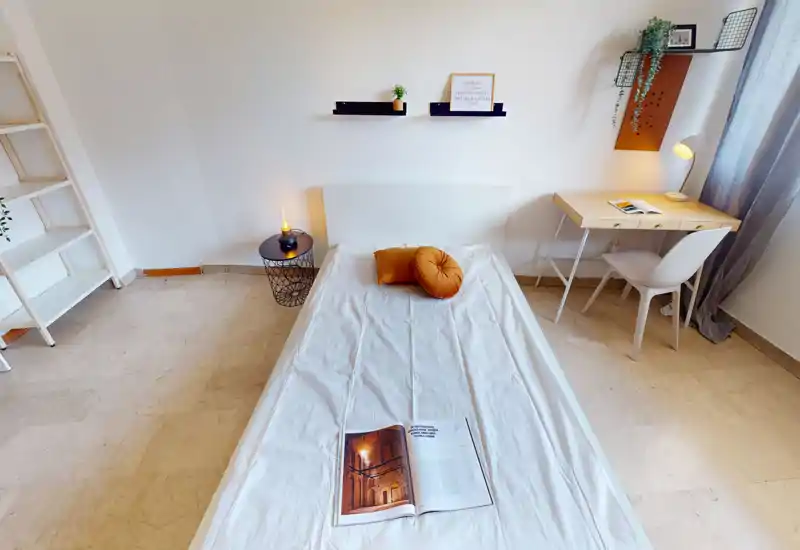 Camera da letto luminosa e minimale con letto singolo al centro, cuscini colorati, comodino con lampada e scrivania vicino alla finestra — ideale per un ambiente in affitto confortevole e ben illuminato.