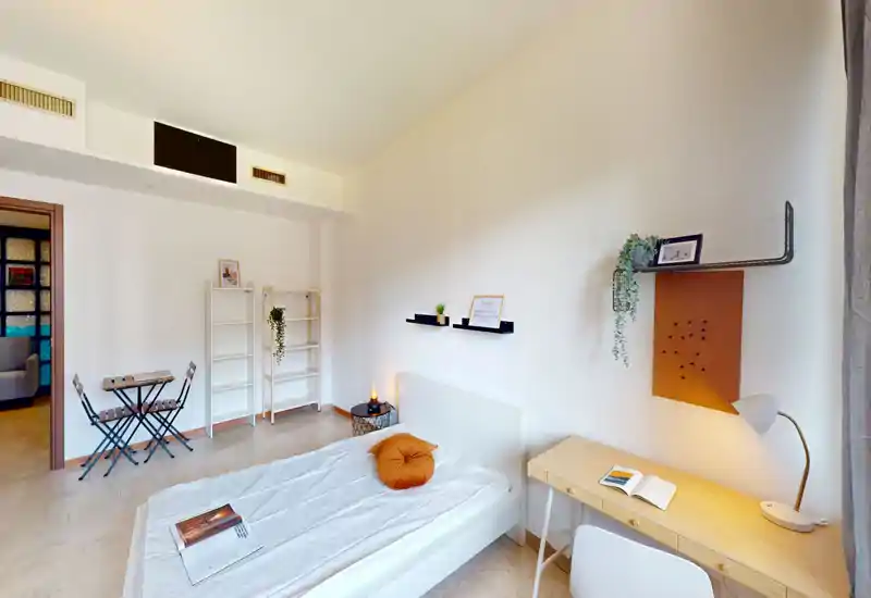 Camera da letto luminosa e minimal con letto bianco, scrivania in legno e semplici dettagli decorativi come un cuscino rotondo e piante, ideale per una persona.