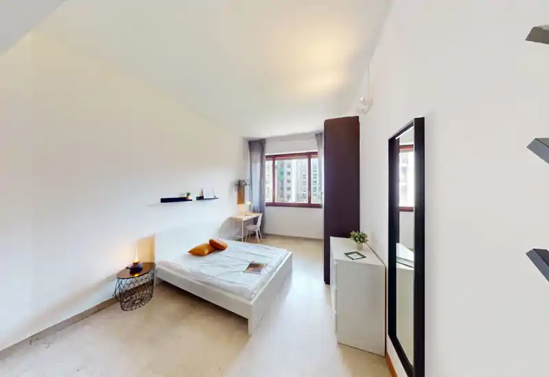 Camera da letto moderna e luminosa con letto matrimoniale al centro, comodino con lampada, scrivania vicino alla finestra e specchio a figura intera. La luce naturale e l’arredamento essenziale creano un’atmosfera calma e accogliente ideale per gli annunci.