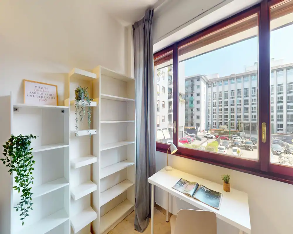 Stanza luminosa e compatta con ampia finestra, una piccola scrivania bianca e scaffalature — ideale come angolo studio o per una camera singola. La luce naturale inonda lo spazio e l'arredamento minimal include piante e una lampada.