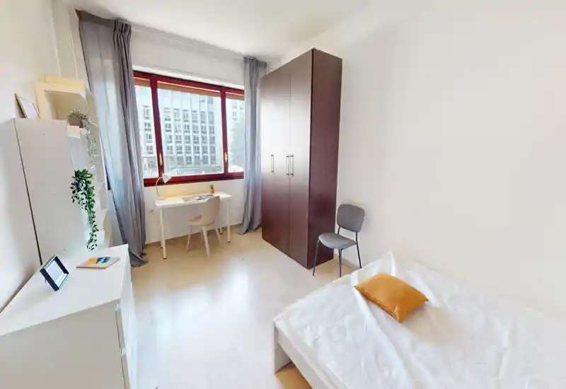 Dormitorio luminoso y minimalista con cama individual, armario, escritorio junto a la ventana y luz natural. La habitación es limpia y funcional con tonos neutros y decoración sencilla.