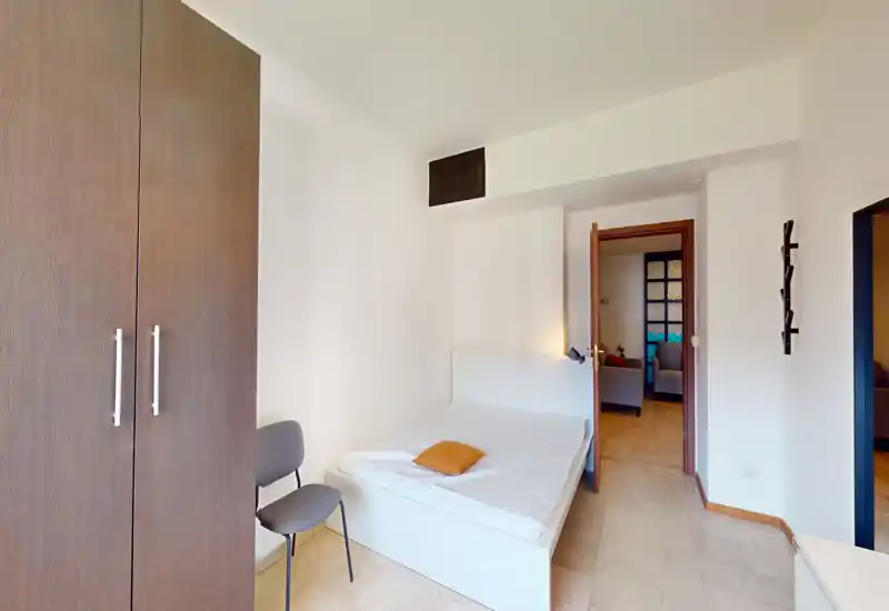 Dormitorio compacto y sencillo con cama individual blanca, armario de madera y suelo de baldosas neutras. La habitación es luminosa y ordenada, ideal para alquileres de corta estancia o estudiantes.