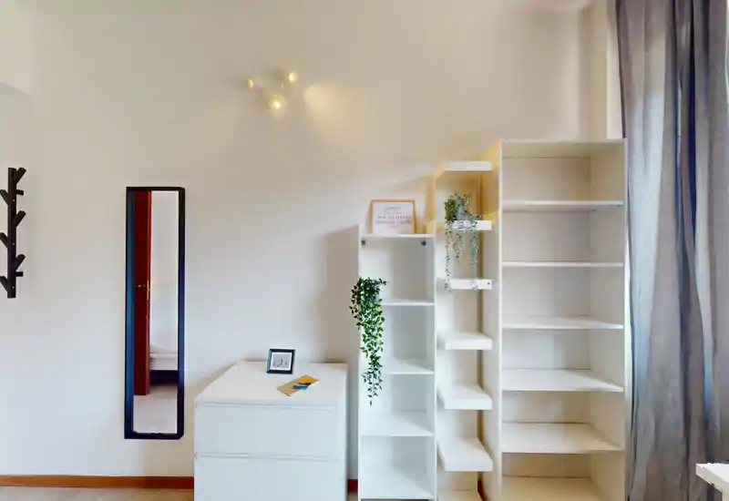 Rincón de dormitorio luminoso y minimalista con estanterías blancas, una cómoda pequeña y un espejo de cuerpo entero; cortinas claras y plantas añaden frescura.