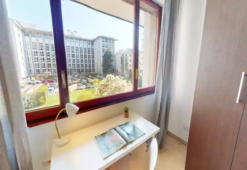 Rincón de habitación luminoso con escritorio blanco junto a una gran ventana, lámpara de mesa y una pequeña planta. La vista da a un patio y edificios vecinos — buena luz natural para leer o trabajar.
