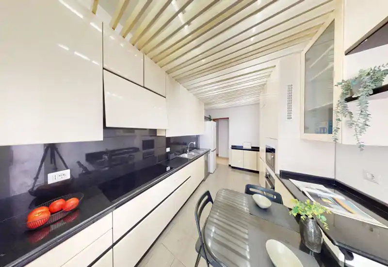Cocina moderna en galera con armarios crema brillantes, encimeras negras y una pequeña mesa; espacio luminoso y ordenado con plantas decorativas y vajilla puesta.