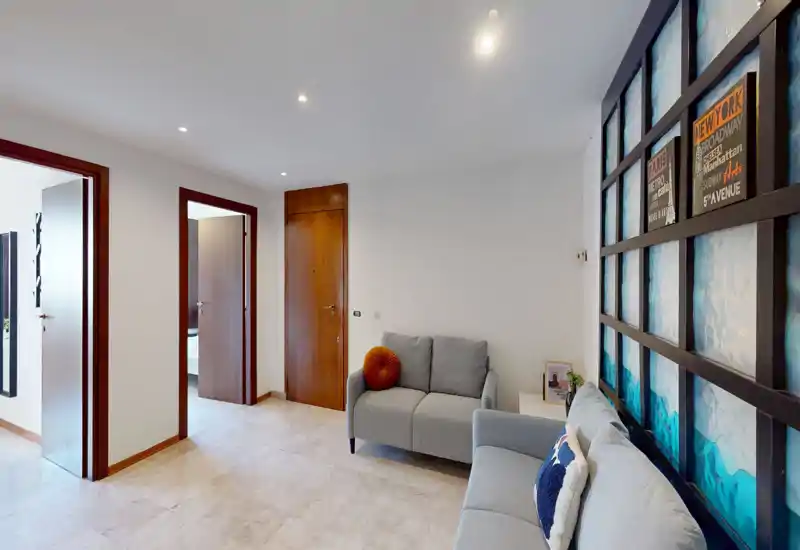 Zona de estar moderna y luminosa con dos sofás grises, cojines decorativos y un gran panel en la pared que crea un espacio acogedor.