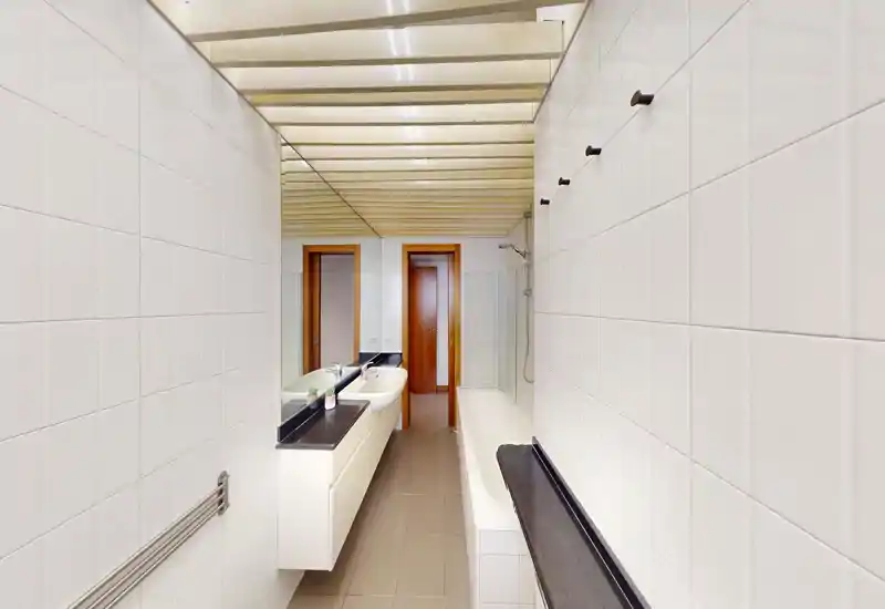 Baño moderno y luminoso con un amplio mueble lavabo, bañera y grifería mural; azulejos blancos y gran espejo que amplían el espacio.