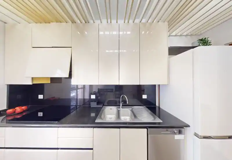 Cocina moderna y compacta con armarios crema brillantes, encimera negra, fregadero y placa integrados, y frigorífico a la derecha — luminosa y ordenada.
