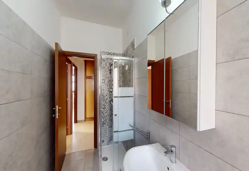 Baño moderno y luminoso con paredes alicatadas, mampara de ducha de cristal, lavabo con espejo-armario y acabados grises neutros.