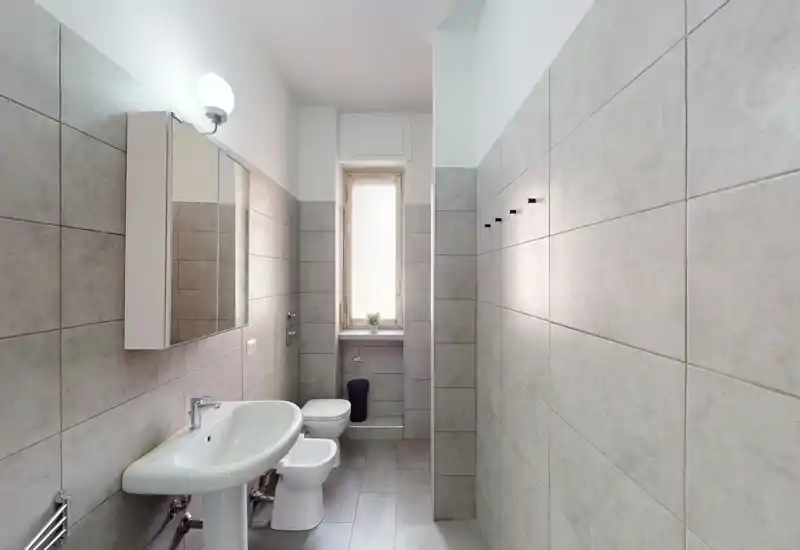 Baño moderno y luminoso con lavabo, bidé, inodoro y armario con espejo sobre el lavabo. Azulejos gris claro en paredes y suelo que crean un aspecto limpio y minimalista, con luz natural de una ventana estrecha.
