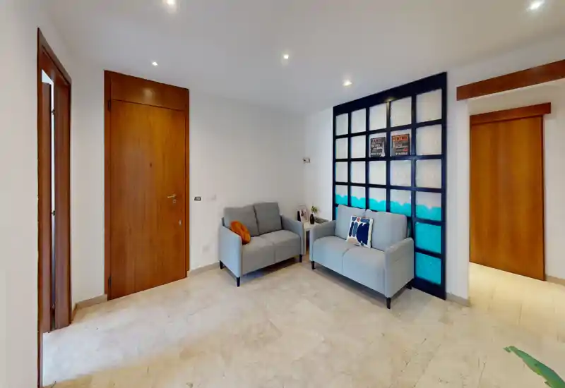 Entrada pequeña y luminosa con dos sofás compactos, separador decorativo y puertas de madera — suelo de mármol y focos empotrados que transmiten una sensación acogedora.