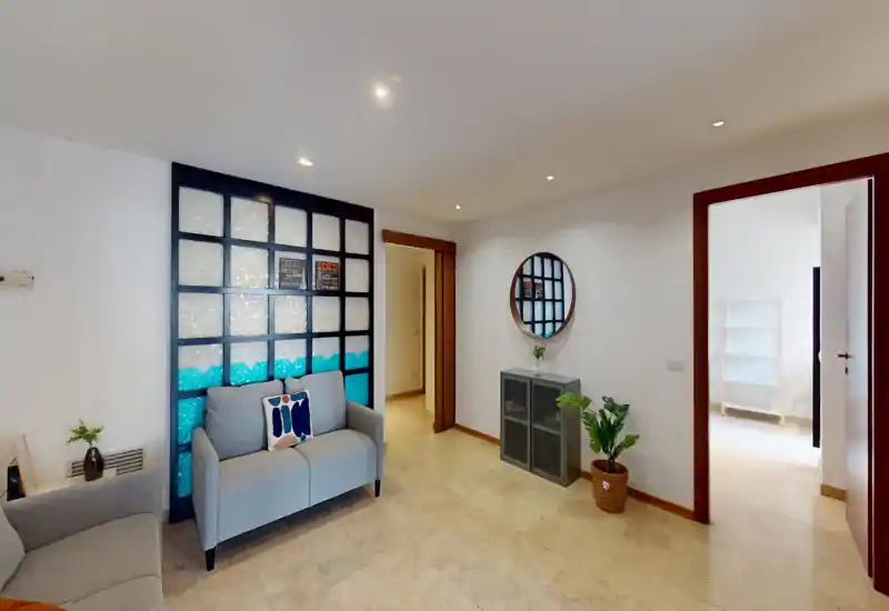 Bright, neatly staged living area with a small sofa, decorative wall panel and mirror; neutral tones and plants create a welcoming atmosphere suitable for a rental listing.