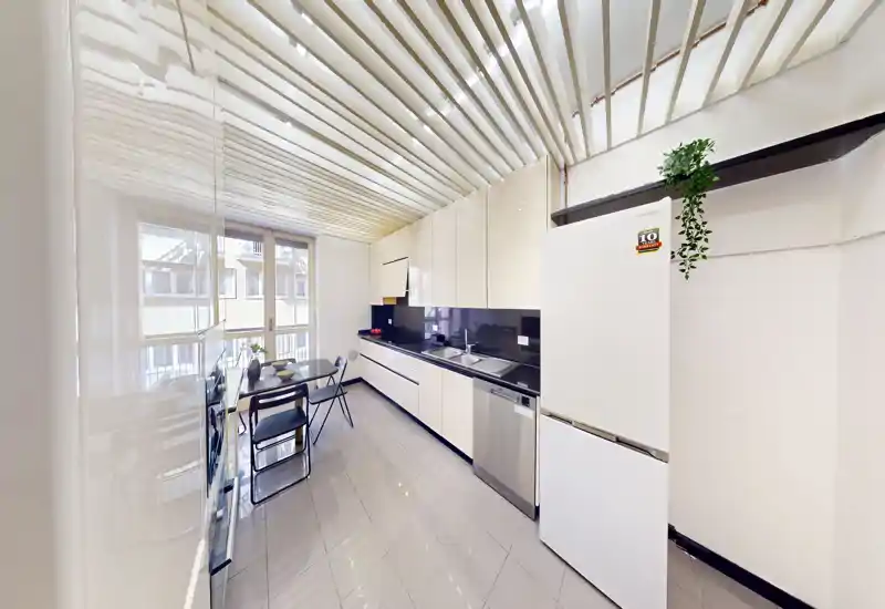 Cocina moderna y luminosa con una larga fila de armarios brillantes, fregadero y lavavajillas integrados, frigorífico independiente y una pequeña mesa junto a la ventana.