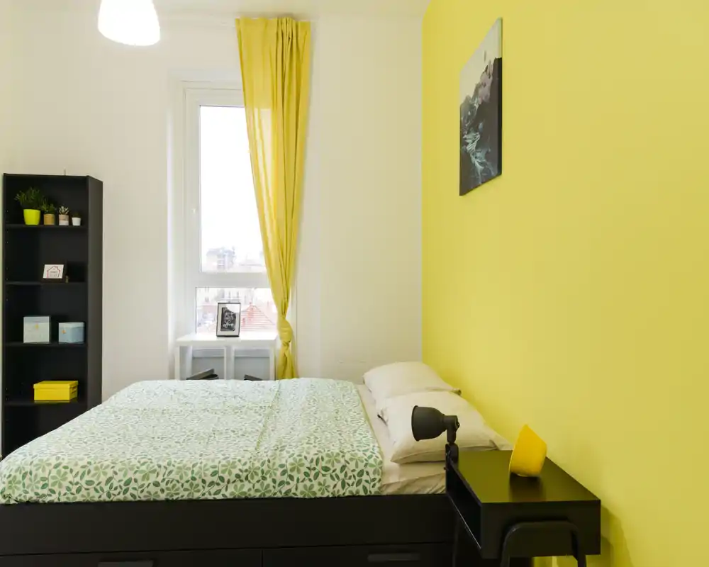 Sunny, modern bedroom with a double bed, patterned green bedding and a bright yellow accent wall. The room features a window with yellow curtains, a bedside table and shelving for storage.